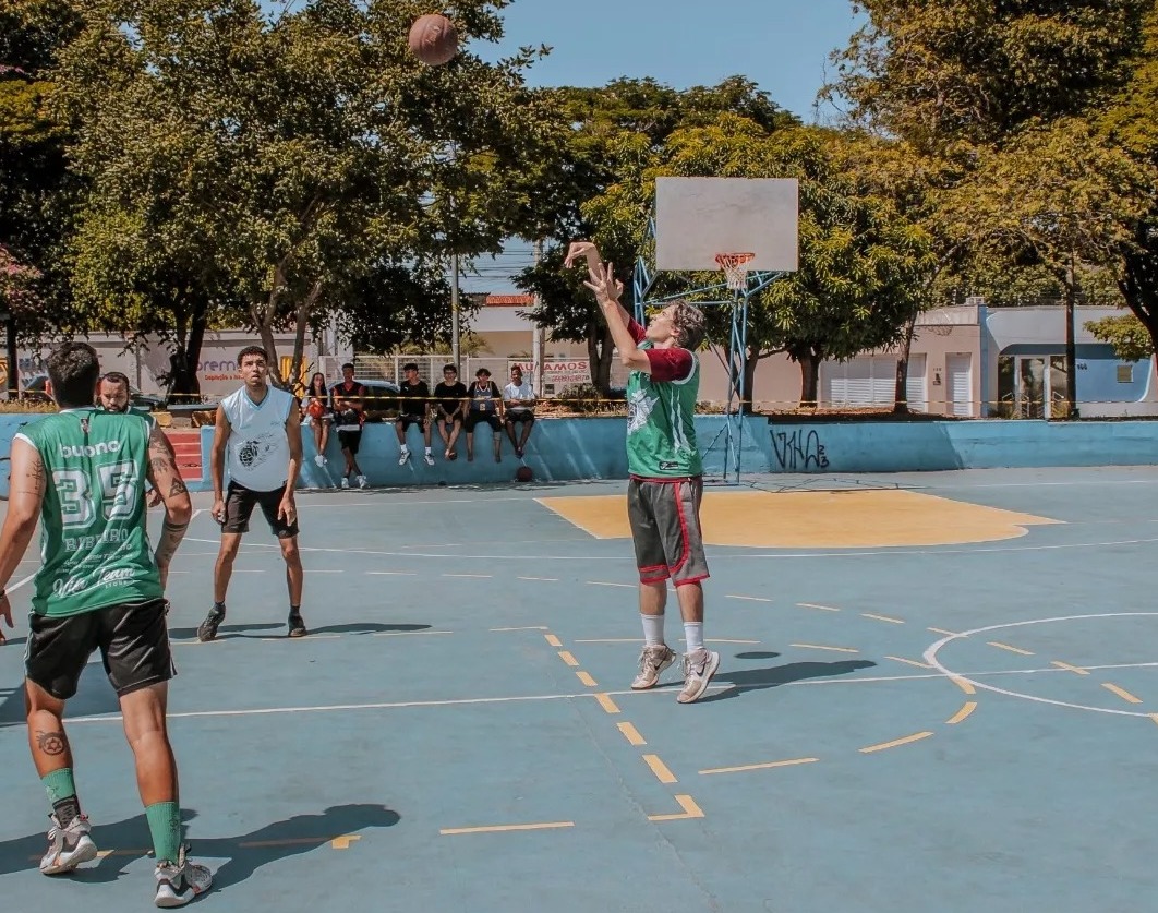 Jogador de basquete arremessando a bola em direção à cesta durante partida na quadra aberta da Praça do Rotary em Montes Claros, com outros atletas observando.
