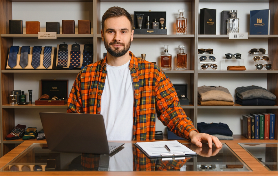 Comerciante mineiro otimista atrás de um balcão com laptop e papéis de venda, diante de prateleiras repletas de produtos masculinos como gravatas, perfumes e óculos.