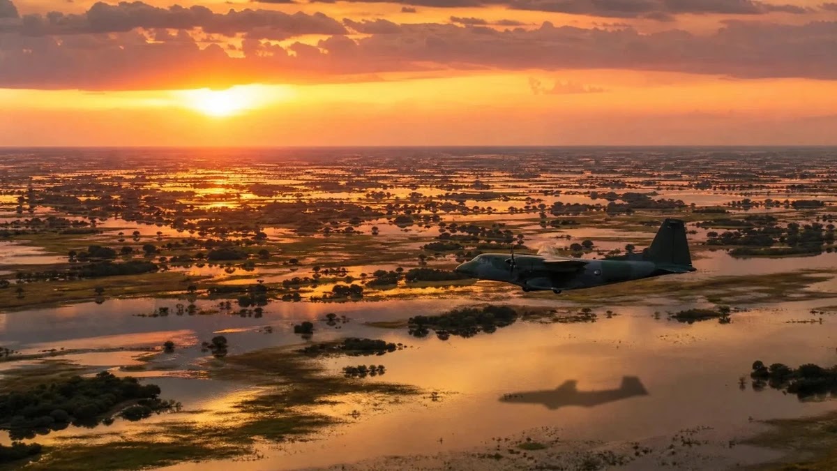 Aeronave militar verde-oliva SC-105 Amazonas da FAB sobrevoando vastidão de lagoas e campos alagados do Pantanal ao pôr do sol em missão de busca.