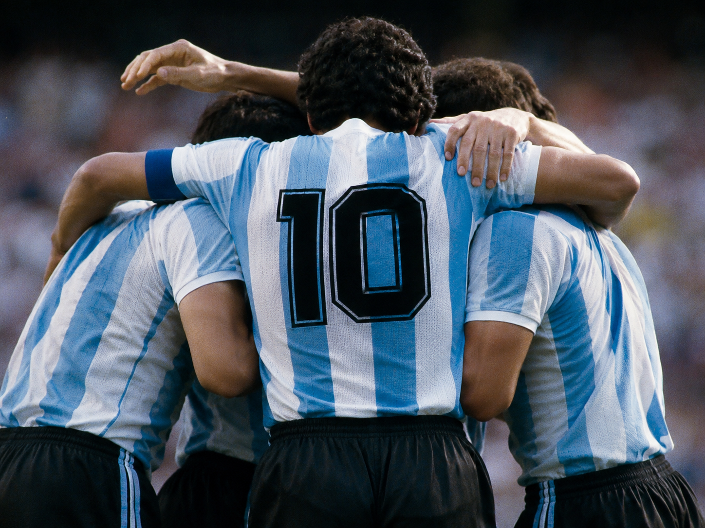 Três jogadores da seleção da Argentina abraçados de costas com a clássica camisa listrada azul e branca, destacando o número 10 centralizado no uniforme retrô.