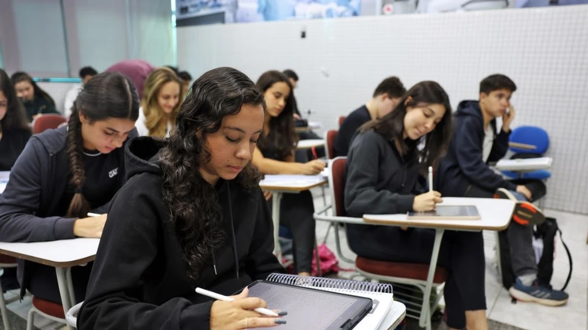 Jovens estudantes sentados em carteiras escolares de uma sala de aula universitária, utilizando tablets e cadernos para anotações.