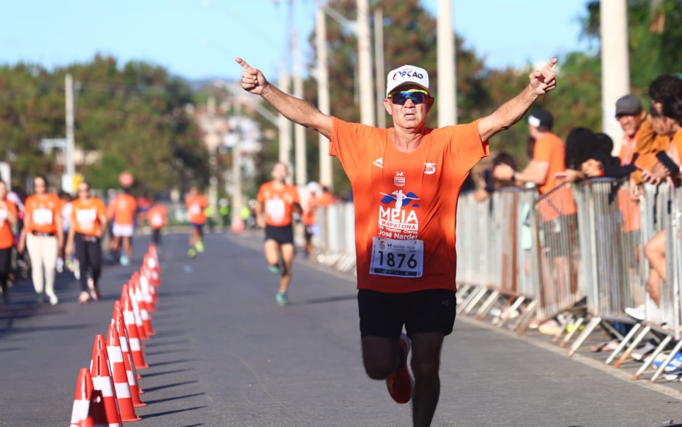 Corredor com camiseta laranja cruzando a linha de chegada de braços abertos na Meia Maratona de Montes Claros José Nardel. Ao fundo, outros corredores e o público na lateral da pista.