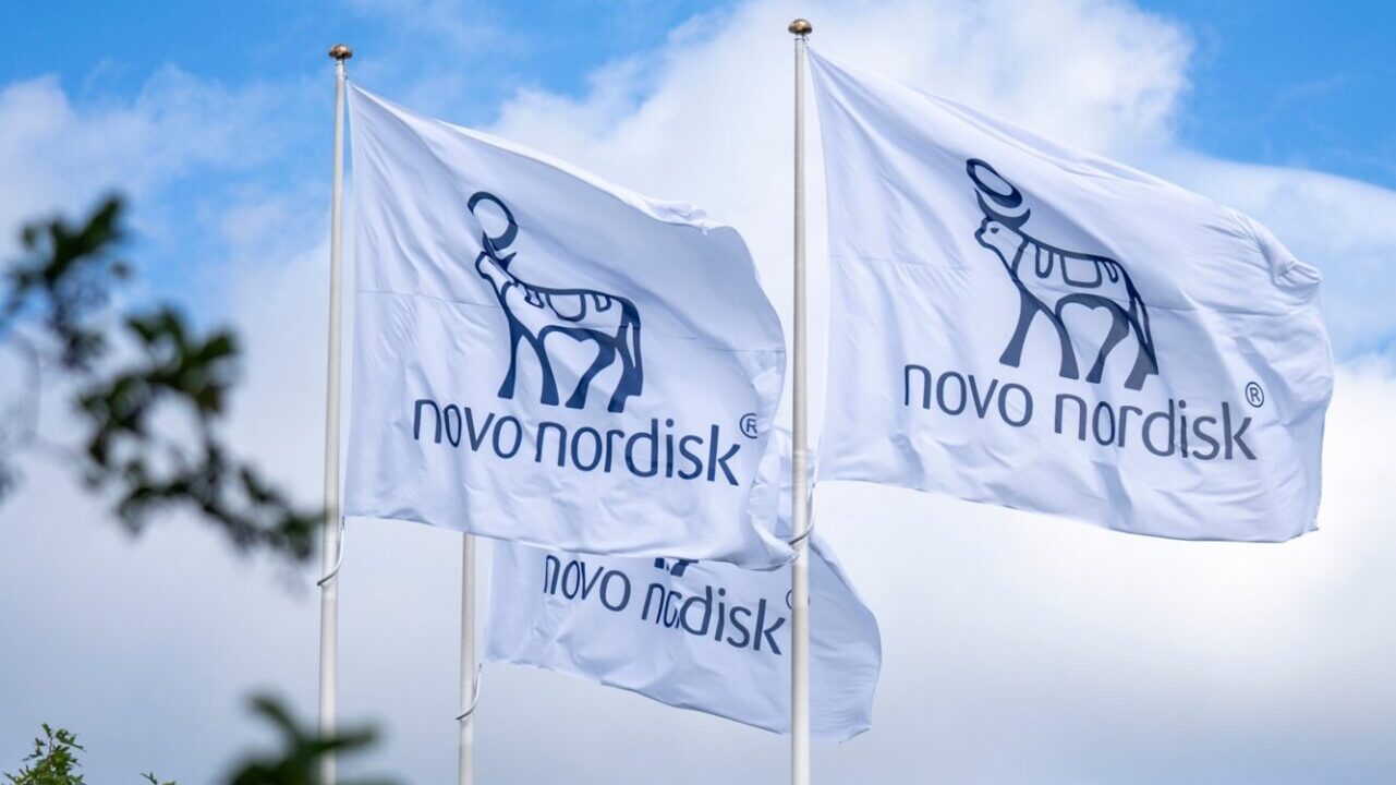 Bandeiras brancas com o logotipo da multinacional farmacêutica Novo Nordisk hasteadas em mastros sob um céu azul com nuvens.