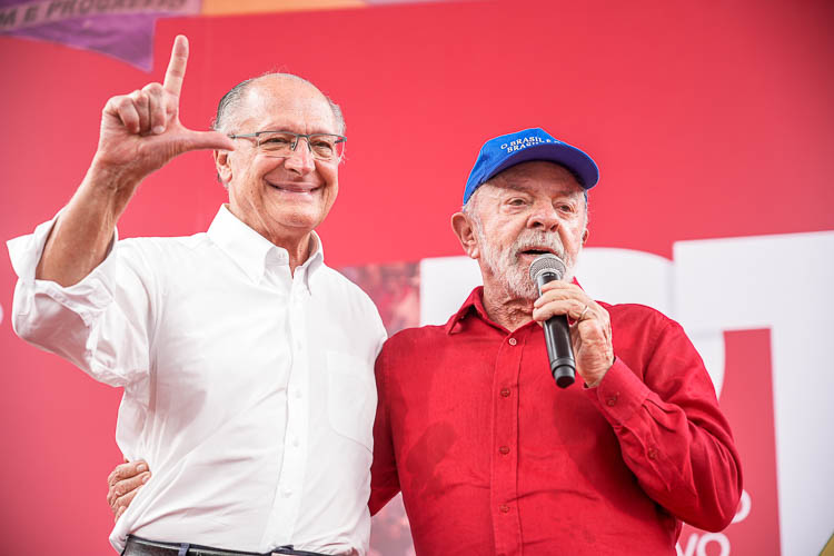Geraldo Alckmin fazendo o sinal com a mão e o presidente Lula discursando com microfone em evento político.