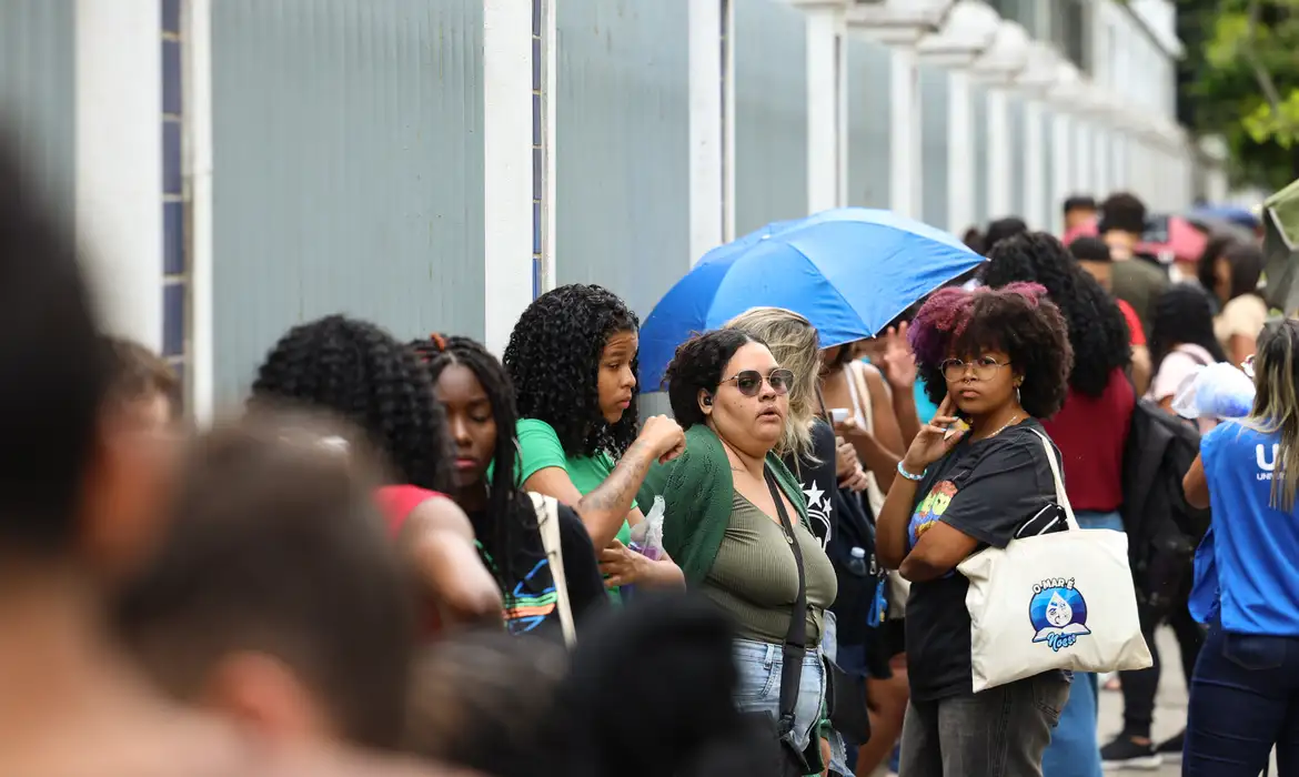 Candidatos do Encceja aguardando em fila na calçada do local de prova, alguns sob guarda-chuva e segurando bolsas.