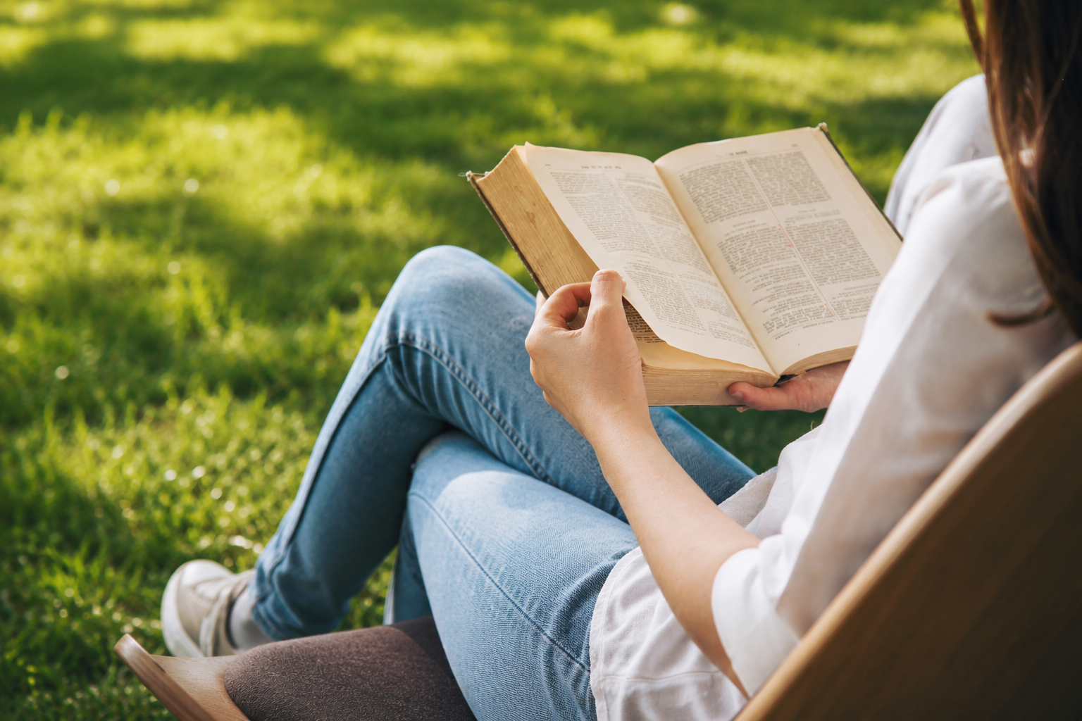 Mulher sentada ao ar livre em uma cadeira de madeira lendo um livro impresso sob a luz do sol.
