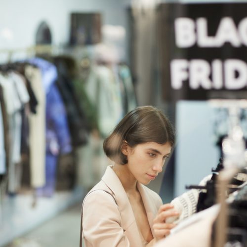 High angle portrait of young woman browsing clothes on rack while shopping in sale season, defocused Black Friday sign in foreground, copy space